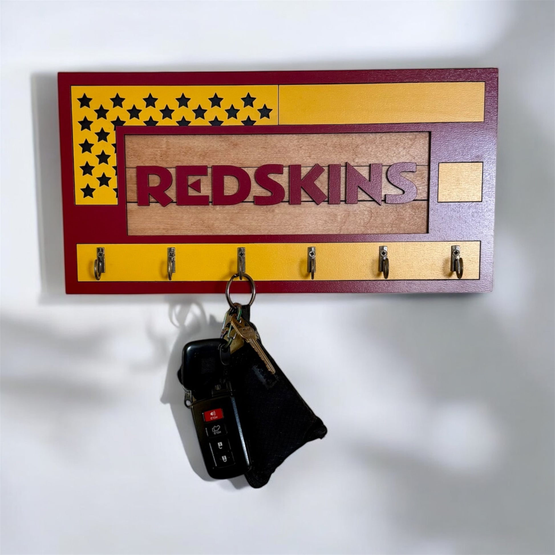 USA flag-inspired wooden key holder with “Redskins” in maroon letters, featuring yellow and maroon accents, six metal hooks, and a rustic maple wood background.